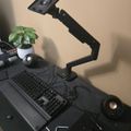 A black desk setup with a mechanical keyboard, monitor arm, small speakers, and a potted plant, arranged on a dark surface against a beige wall.