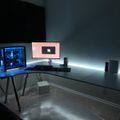 A modern gaming setup with an L-shaped glass desk featuring a blue-lit PC tower, curved monitor, keyboard, and speakers in a dimly lit room with LED strip lighting under the desk