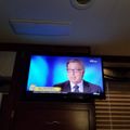 Television screen showing a news broadcast with a male anchor in a suit and glasses against a blue background, mounted on a wall in what appears to be an RV or mobile home interior