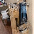 A wall-mounted electrical outlet box under construction, wrapped in clear plastic sheeting with exposed wiring and black electrical components visible inside the tan-colored outlet box installed in a beige textured wall.