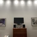 A room with white walls featuring track lighting, a wooden dresser with a TV and speakers on top, and two framed artworks hanging on either side of the entertainment setup.