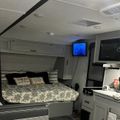Interior of a modern RV or travel trailer showing a bed area with patterned bedding, kitchen counter with appliances, mounted TV screen, and white cabinetry throughout the compact living space