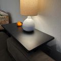 A table lamp with white ceramic base and beige fabric shade sits on a dark wooden surface next to a small orange decorative object