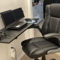 A home office setup with a black leather office chair positioned at a dark wooden desk that holds a laptop, external monitor, and various cables, with white kitchen cabinets visible in the background