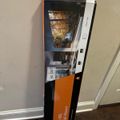A large flat-screen TV box leaning against a white wall on a dark wooden floor, with orange and black packaging displaying product images
