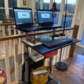 A standing desk setup with two laptops, an external keyboard, and a desk lamp in a bright room with large windows and wooden railings