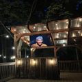 Outdoor wooden pergola structure with string lights and a large TV screen displaying a person, set on a wooden deck at night surrounded by trees