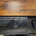 A black gaming keyboard and mouse with blue LED backlighting positioned on a dark mouse pad, placed on a wooden desk surface