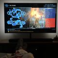 A gaming setup showing a large TV screen displaying a video game called Meltdown with a scoreboard and game interface, with a couch and gaming controllers visible in the foreground