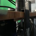 A desk setup with a monitor arm mounted to a wooden desk surface, with cables and equipment visible against a green screen background