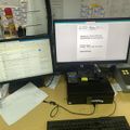 A dual monitor computer workstation setup on a desk showing documents and forms, with a keyboard, mouse, phone, and various office supplies visible in a busy office environment
