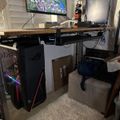 A computer gaming setup with a standing desk, PC tower with RGB lighting, monitor displaying a game, and gaming peripherals in a home office space.
