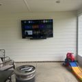 A covered patio or sunroom with white shiplap walls, featuring a wall-mounted TV displaying a streaming service menu, outdoor furniture including chairs and a round table, children's toys including a colorful slide and ride-on truck, and concrete flooring.