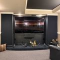 A home theater room with dark walls, a large wall-mounted TV displaying a movie scene, decorative dragon figurines on a shelf below the screen, and comfortable seating with a patterned area rug.