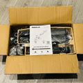 An open cardboard box containing a dual monitor desk mount with instruction manual and various black components wrapped in plastic packaging