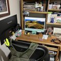 A home office workspace with two computer monitors on a wooden desk, an office chair, and wooden shelving filled with books and papers