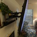 A home office setup with a curved monitor on an adjustable arm mount positioned above a fireplace mantel, with a chair covered by a patterned blanket nearby and a plant visible on the left side.