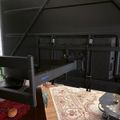 A home office setup with multiple monitors mounted on an adjustable arm above a desk, with decorative rugs on hardwood flooring and dark ceiling beams visible above.