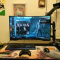 A computer gaming setup with monitor displaying a video game character selection screen, keyboard, mouse, and controller on a desk with various decorations on the wall