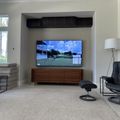 Modern living room with wall-mounted TV displaying a golf scene, wooden media console, black leather chair with ottoman, and floor lamp near a window