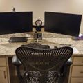 A dual monitor computer workstation setup on a granite countertop with a black ergonomic office chair, keyboard, and various desk accessories including a webcam and calculator