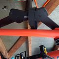 Orange bicycle frame with black handlebars and components in what appears to be a garage or storage area with wooden beams visible in the background
