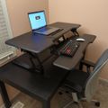 A home office setup with a black adjustable standing desk converter placed on a dark wooden desk, featuring a laptop, keyboard, and mouse, with an ergonomic office chair positioned in front