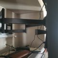 A desk setup with a black monitor arm mounted above a shelf containing books and various cables, with white walls in the background