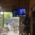 A garage or workshop interior with a mounted TV displaying NFL content, tools and equipment visible, and an open door showing an outdoor patio area with construction materials