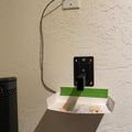 A wall-mounted device with a curved cable connecting to a white mounting plate above, with a black component and green tape visible on a white shelf below