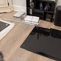 A home office setup on a wooden floor showing furniture assembly in progress, with black panels, white packaging materials, rolled items, a black shelving unit with decorative items, office equipment including a printer and trash bin, and cream-colored cabinetry visible in the background.