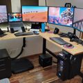 A modern home office setup with multiple monitors displaying scenic landscapes, an L-shaped wooden desk with black legs, ergonomic office chair, computer tower, and various tech accessories in a room with hardwood flooring and white walls.