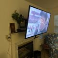 Living room with mounted TV above fireplace showing a movie scene, floral armchair, and home decor