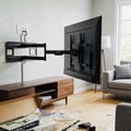 A modern living room with a wooden TV stand, wall-mounted flat-screen TV with articulating arm mount, gray upholstered armchair, and hardwood flooring. Installation materials and tools are visible on the floor in the foreground.