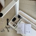 Partially assembled white desk or table frame with instruction manual, tools, and hardware scattered on beige carpet during furniture assembly process