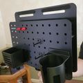 A black pegboard organizer with mounting slots and holes, featuring attached storage containers and hooks for organizing tools or supplies
