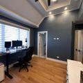 Modern home office with dark blue accent wall, white coffered ceiling with ceiling fan, hardwood floors, black desk with dual monitors, and white trim throughout