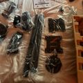 Unboxed computer monitor components and hardware parts laid out on a beige surface, including various black plastic brackets, cables, and mounting accessories in clear plastic packaging, with an instruction manual visible at the top.