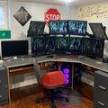 A professional gaming and computer workstation setup featuring a curved desk with multiple monitors displaying matrix-style code, a red gaming chair, purple LED lighting in the PC case, audio equipment, and various decorative items including street signs and posters on the walls.