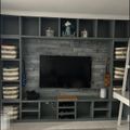 Built-in entertainment center with gray stone fireplace surround, flat screen TV, shelving units with storage baskets, and decorative items including a small guitar