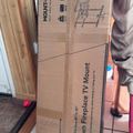 A cardboard box for a pull-down fireplace TV mount leaning against wooden boards in what appears to be a storage or construction area