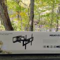 A TV wall mount product box displayed outdoors on a wooden surface with trees and foliage visible in the background