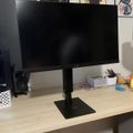 A computer monitor on a stand sits on a wooden desk with speakers, storage containers, and anime-style posters visible on the wall behind it