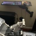 A workspace showing a white microscope or scientific instrument in the foreground with a keyboard mounted on an adjustable arm above it, and computer monitors visible in the background