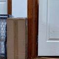 A cardboard package sits on a wooden floor next to a white door with brown wooden door frame, with a wire basket visible on the left side
