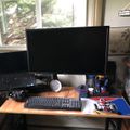 A computer workstation with a large monitor, keyboard, and various items on a wooden desk near a window with natural lighting