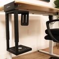A height-adjustable standing desk with wooden top and black metal frame, featuring an under-desk computer tower mount, positioned next to an office chair on a jute rug
