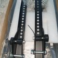 Two black metal brackets with multiple holes and adjustment mechanisms, packed in white foam padding inside a cardboard box