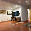 A modern living room with polished concrete flooring, featuring a wall-mounted flat-screen TV above a two-tone wooden media console. The space includes recessed lighting, framed artwork on white walls, and opens to an adjacent room with natural light from windows and glass doors.