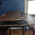 A wooden cutting board sits on top of a black laptop computer on a desk near a window with blue walls in the background